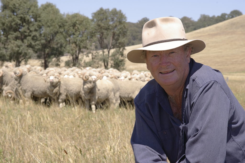 Photo of farmer in hat and blue shirt with sheep behind him. 